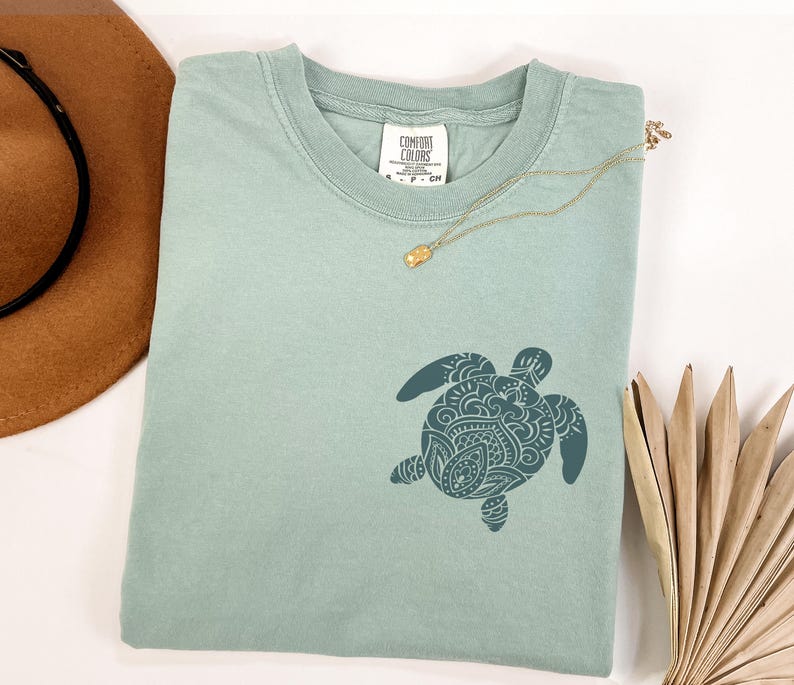 May include: A light green Comfort Colors t-shirt featuring a detailed teal turtle design. A gold necklace with a pendant rests on the shirt. A brown hat and dried palm leaves are in the background.