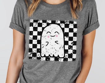 May include: A grey t-shirt with a smiling ghost graphic. The ghost is white with pink cheeks and black eyes, set against a black and white chequered background. The shirt has rolled-up sleeves.