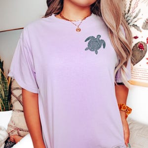 May include: A light purple t-shirt featuring a detailed turtle design on the front. The short-sleeved shirt is made of soft material and has a relaxed fit. The turtle design is a dark teal color.