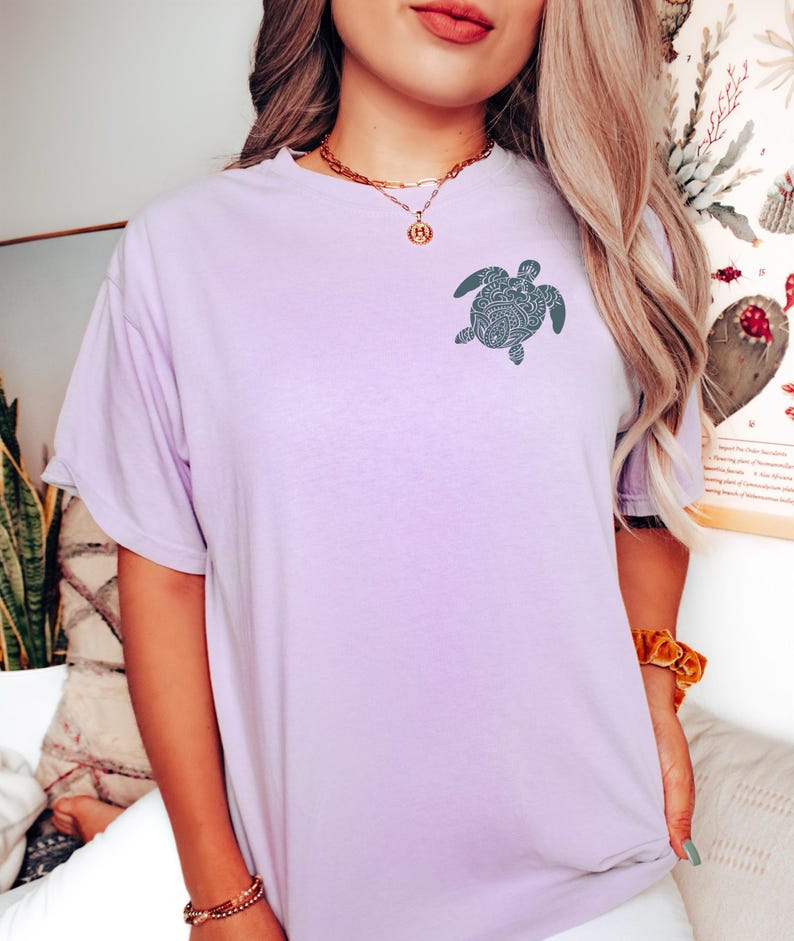 May include: A light purple t-shirt featuring a detailed turtle design on the front. The short-sleeved shirt is made of soft material and has a relaxed fit. The turtle design is a dark teal color.