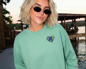 May include: A sage green crewneck sweatshirt featuring a small embroidered graphic on the left chest. The design is a heart-shaped depiction of the Earth, showcasing blue oceans and green landmasses with a dark outline, set against the solid fabric.