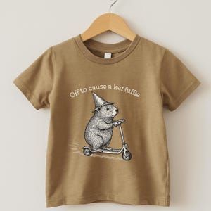 May include: Tan short-sleeved t-shirt with a brown collar. The shirt features a black and white illustration of a rodent wearing a wizard hat and riding a scooter. The text "Off to cause a kerfuffle" is above the illustration.