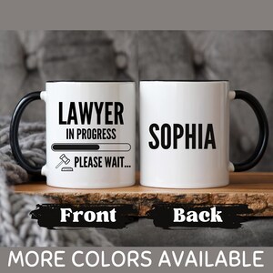 May include: White ceramic mug with a black handle and rim. The front of the mug says "LAWYER IN PROGRESS" with a progress bar and gavel icon. The back of the mug has the name "SOPHIA" in bold black letters. The bottom of the image says "MORE COLOURS AVAILABLE".