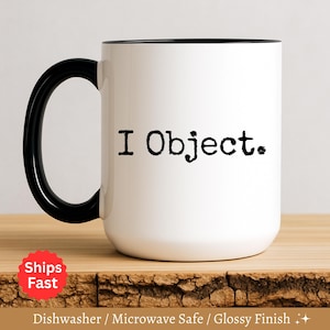 May include: White ceramic mug with a black handle and rim. The mug features the text "I Object" in black, centered on the front. The mug is on a wooden surface.