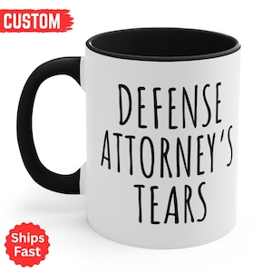 May include: White ceramic mug with a black handle and interior. The mug features the text "DEFENSE ATTORNEY'S TEARS" in black, bold letters. A red "CUSTOM" label is in the upper left, and a red "Ships Fast" label is in the lower left.