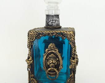 May include: A blue glass decanter with a gold metal frame and a lion head design. The decanter has a clear glass stopper.