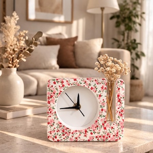 May include: A square clock with a white face and black hands, surrounded by a floral pattern of red and pink flowers on a white background. A small glass vase with dried flowers is attached to the side. The clock is on a light-colored surface.