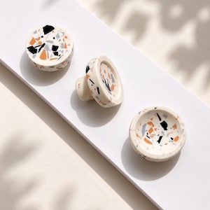 May include: Three round cabinet knobs featuring a cream-coloured base with a multi-coloured terrazzo pattern. The speckled design includes small, irregular fragments in shades of black, light blue, and orange, distributed across the smooth surface.