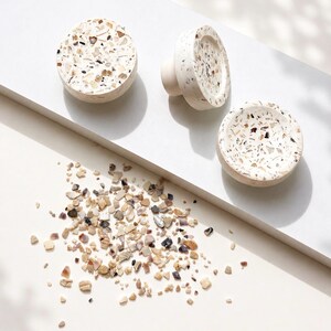 Op de afbeelding: Drie ronde kastknoppen met een witte basis ingelegd met veelkleurige steen- en schelpfragmenten. De knoppen hebben een glad, afgerond profiel met een platte voorkant en een uitstekende cilindrische basis voor bevestigingsmateriaal.