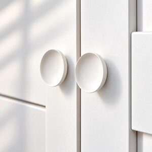 May include: Two circular, matte white cabinet knobs are mounted on a white cabinet door. The knobs have a concave, bowl-like centre and a smooth, rounded edge, providing a simple, functional grip for opening the cabinet doors in a kitchen or room.