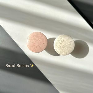 Pode incluir: Dois objetos redondos e texturizados, um rosa claro e outro branco sujo, estão sobre uma superfície branca. O texto "Sand Series" está presente. O fundo apresenta sombras e um elemento diagonal cinza escuro.