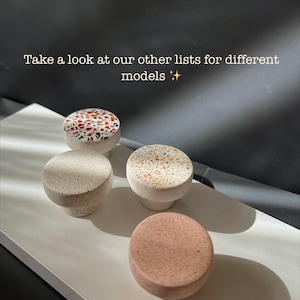 Op de afbeelding: Vier decoratieve kastknoppen in verschillende ontwerpen. De knoppen hebben een rood-wit terrazzo patroon, een gespikkeld beige en een effen roze. De tekst "Take a look at our other lists for different models" is zichtbaar.