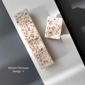 Könnte beinhalten: Zwei rechteckige Dekorationsstücke mit einem Mokka-Terrazzo-Design. Das größere Stück ist lang und schmal, während das kleinere quadratisch ist. Die Stücke haben eine cremefarbene Basis mit braunen und orangefarbenen Flecken. Der Text "Mocha Terrazzo Design" ist sichtbar.