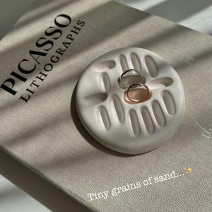 May include: A white, circular ring holder with oval cutouts, holding a silver and a rose gold ring. The holder rests on a book with the title "PICASSO LITHOGRAPHS". The text "Tiny grains of sand..." is at the bottom.