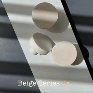 Op de afbeelding: Drie beige ronde decoratieve knoppen en handgrepen. Eén knop heeft een paddenstoelvormig ontwerp. De tekst "Beige Series" is zichtbaar. De knoppen zijn gerangschikt op een wit en grijs oppervlak.