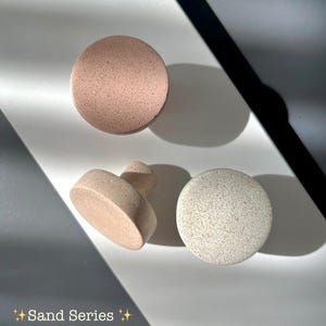 Puede incluir: Tres piezas de cerámica en tonos neutros. Dos son redondas y una tiene un asa. El texto "Sand Series" se encuentra en la parte inferior izquierda de la imagen.