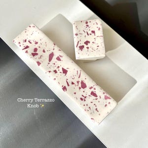 May include: Two cherry terrazzo knobs with a rectangular shape and a white base. The knobs have a pattern of red and pink flecks. The text "Cherry Terrazzo Knob" is visible on the image.