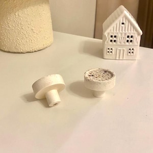 May include: A cream-coloured table lamp with a textured base and a neutral lampshade. Two ceramic cabinet knobs are in the foreground. A small white ceramic house with windows and a door is also present.