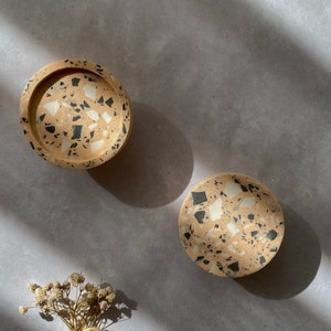 May include: Two round terrazzo coasters in beige with black and white flecks. The coasters are on a grey surface with a bunch of dried flowers.