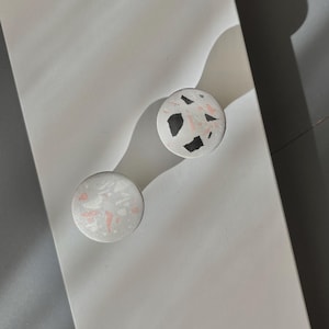 Pode incluir: Dois puxadores redondos com um design salpicado de preto, rosa e branco sobre um fundo cinza claro. Os puxadores são feitos de um material sólido, provavelmente para móveis ou decoração.