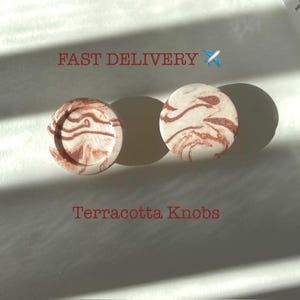 May include: Two round terracotta knobs with a white and brown marbled design.  "Fast Delivery" and a plane emoji are above the knobs.  "Terracotta Knobs" is written below the knobs.