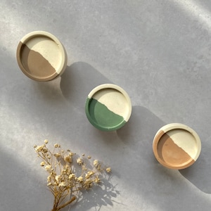 Peut inclure: Trois sous-verres ronds en béton avec une base blanche et une partie supérieure colorée. Les sous-verres sont dans des tons de brun, vert et beige. Un petit bouquet de fleurs blanches séchées se trouve au centre de l'image.