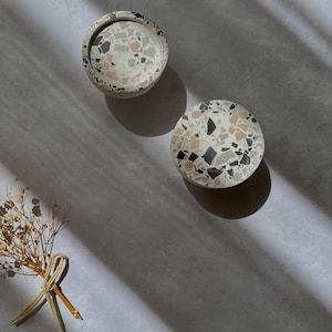 May include: Two round terrazzo coasters in white, grey, and black with a small bouquet of dried white flowers on a grey background.