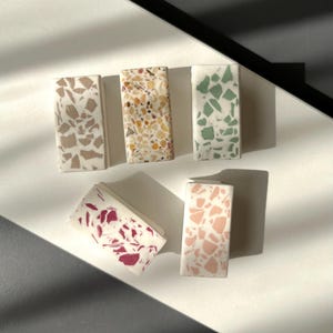 Puede incluir: Una colección de baldosas decorativas rectangulares con patrones de terrazo. Las baldosas presentan varios colores, incluyendo beige, amarillo, verde, rosa y burdeos, sobre un fondo blanco. Las baldosas están dispuestas sobre una superficie blanca y gris.