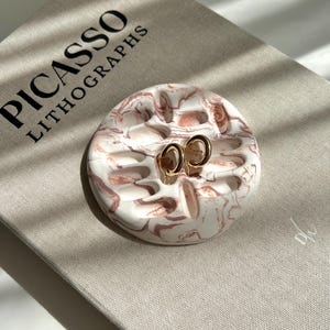 May include: A pair of gold hoop earrings rests on a white and pink marbled ceramic dish. The dish is on top of a book titled "PICASSO LITHOGRAPHS". The earrings are small and circular, and the dish has a unique, organic shape.