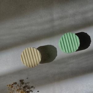 May include: Two round, textured soap bars, one green and one beige, on a white surface with a dried flower sprig.