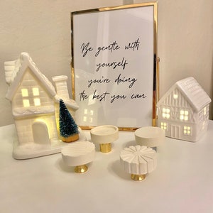 May include: Five white ceramic knobs with gold accents. The knobs are in various shapes and sizes. The knobs are on a white surface with a framed print that reads "Be gentle with yourself you're doing the best you can".