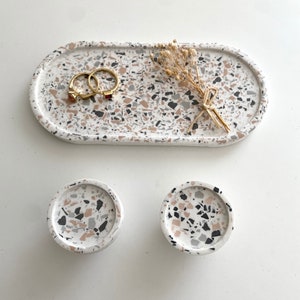 Może przedstawiać: Zestaw trzech tacek z terazzo w kolorze białym, czarnym i różowym. Tace mają kształt owalny i okrągły. Tace idealnie nadają się do przechowywania biżuterii, kluczy lub innych drobnych przedmiotów.