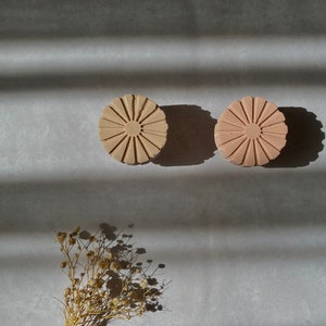May include: Two round, light brown, decorative soap dishes with a daisy-like design. The dishes are on a gray surface with white stripes. A small bouquet of dried flowers is in the lower left corner of the image.