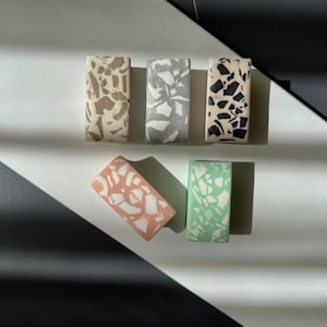 Pode incluir: Seis objetos decorativos retangulares em estilo terrazzo em várias cores. Os objetos apresentam um padrão salpicado com cores como bege, cinza, preto, pêssego e verde. Os objetos são dispostos em uma superfície branca com detalhes geométricos em preto e branco.