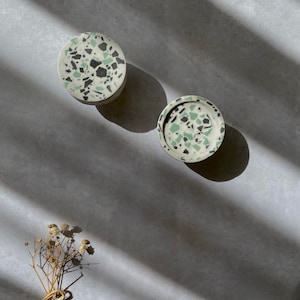 May include: Two round coasters with a terrazzo design. The coasters feature a light background with green, black, and white flecks. The coasters are made of a solid material and are sitting on a gray surface.