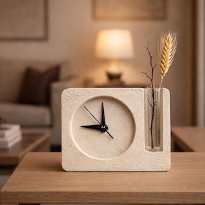 Puede incluir: Un reloj cuadrado color crema con manecillas negras y un pequeño jarrón. El jarrón contiene una ramita de trigo y una pequeña rama. El reloj está sobre una mesa de madera.