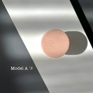 Puede incluir: Una esponja de maquillaje redonda, de color rosa claro, con una superficie texturizada. La esponja est&aacute; etiquetada como "Model A" en texto blanco. El fondo es una mezcla de blanco, negro y gris, con sombras.