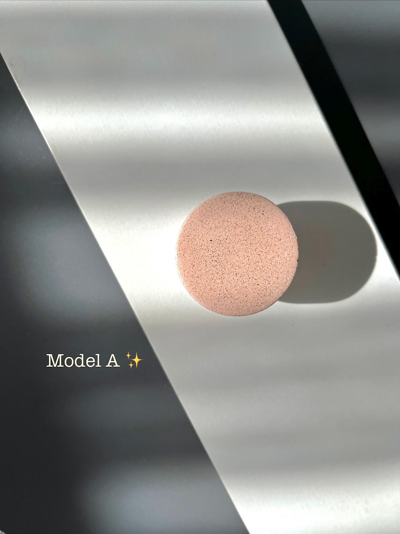 Puede incluir: Una esponja de maquillaje redonda, de color rosa claro, con una superficie texturizada. La esponja est&aacute; etiquetada como "Model A" en texto blanco. El fondo es una mezcla de blanco, negro y gris, con sombras.