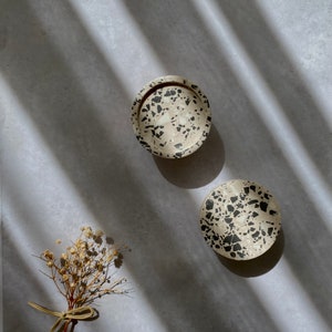 Pode incluir: Dois porta-copos redondos de terrazzo com fundo branco e manchas pretas. Os porta-copos estão sobre uma superfície cinza claro com sombras de uma janela.