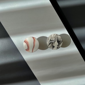 Pode incluir: Dois brincos redondos. Um é branco com veios vermelhos, o outro é marmoreado em preto e branco. Os brincos são exibidos em uma superfície prateada com uma sombra diagonal.