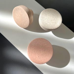 Puede incluir: Tres pomos de armario redondos y moteados en tonos beige y rosa. Un pomo tiene un asa redondeada, mientras que los otros son esferas y discos simples. Los pomos est&aacute;n dispuestos sobre una superficie blanca con un fondo oscuro.