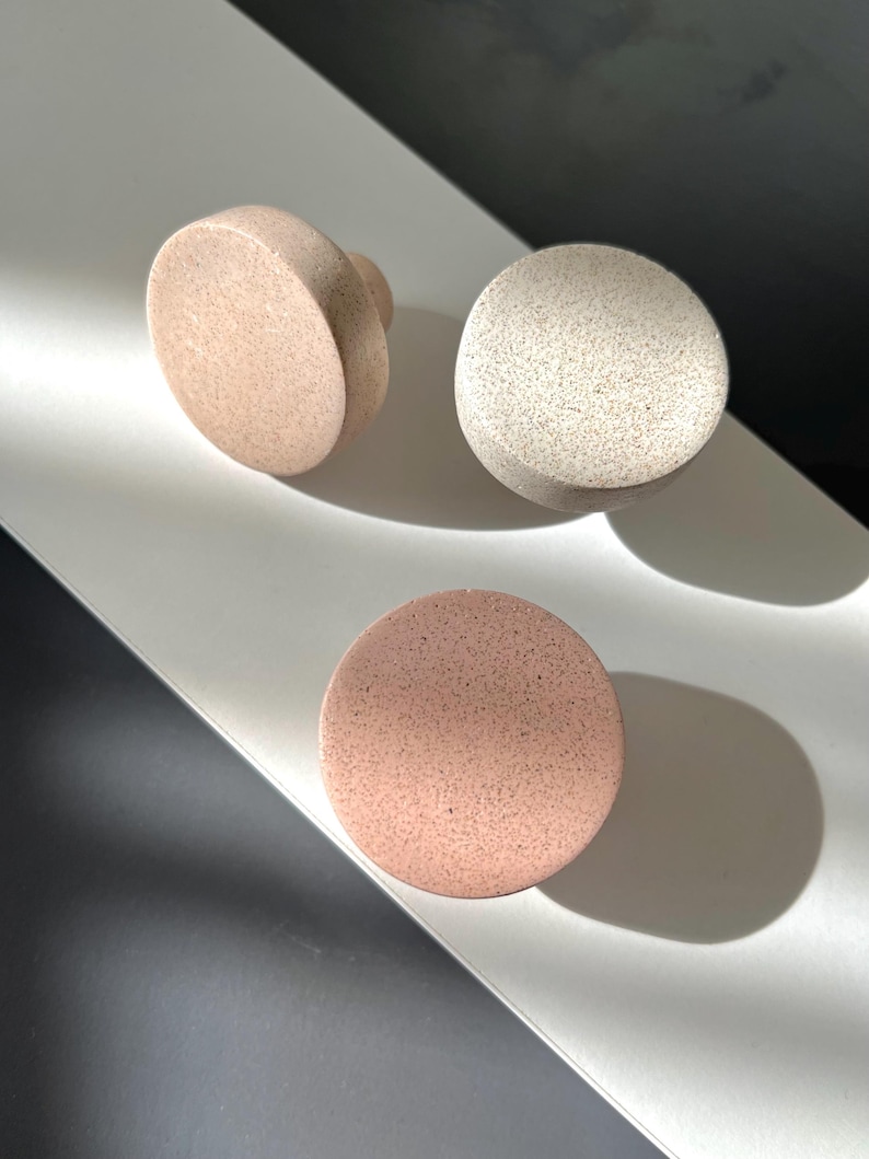 Puede incluir: Tres pomos de armario redondos y moteados en tonos beige y rosa. Un pomo tiene un asa redondeada, mientras que los otros son esferas y discos simples. Los pomos est&aacute;n dispuestos sobre una superficie blanca con un fondo oscuro.