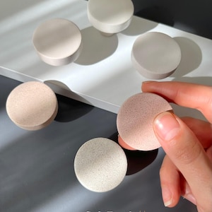 Puede incluir: Un surtido de pomos redondos y esponjas cosm&eacute;ticas en tonos neutros. Los pomos son en tonos blanco, beige y gris. Una esponja es sostenida por una mano, y las otras est&aacute;n dispuestas sobre una superficie blanca. El texto "Soft Touch" es visible.