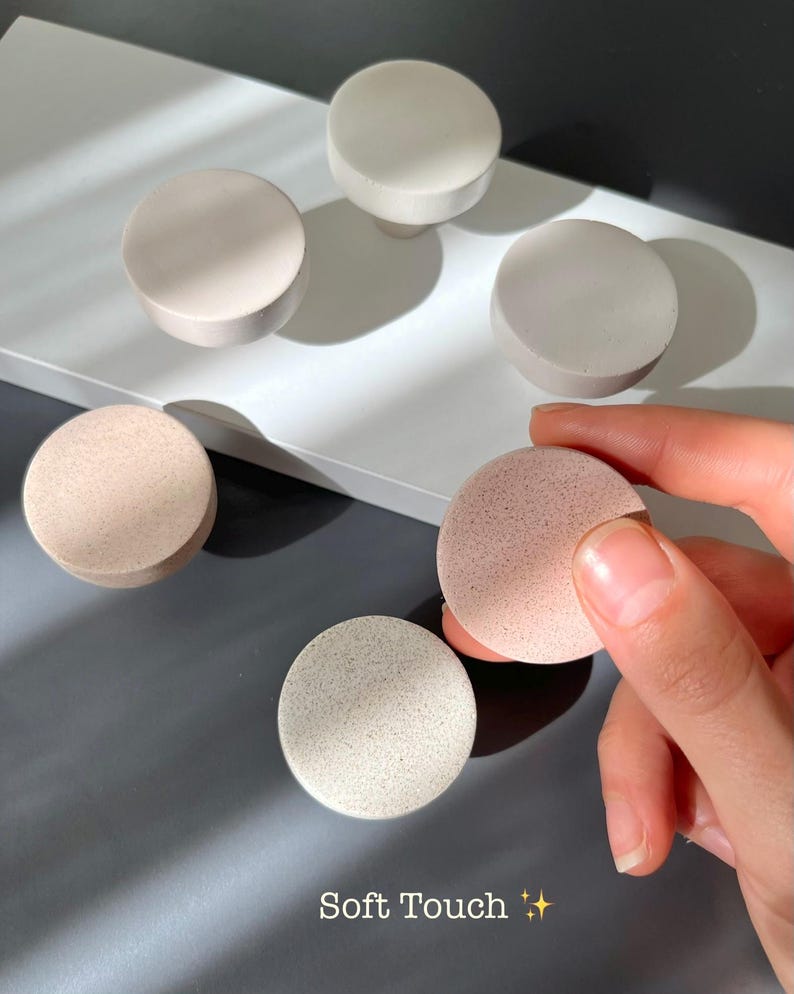 Puede incluir: Un surtido de pomos redondos y esponjas cosm&eacute;ticas en tonos neutros. Los pomos son en tonos blanco, beige y gris. Una esponja es sostenida por una mano, y las otras est&aacute;n dispuestas sobre una superficie blanca. El texto "Soft Touch" es visible.