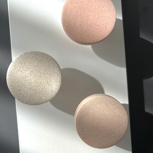 Puede incluir: Tres objetos decorativos redondos y moteados en tonos rosa y beige. Est&aacute;n dispuestos sobre una superficie blanca, con sombras que a&ntilde;aden profundidad. La textura moteada y los colores neutros sugieren un dise&ntilde;o minimalista.