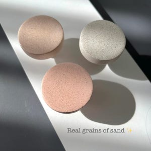 Puede incluir: Tres objetos redondos y texturizados en tonos rosa y gris se disponen sobre una superficie blanca. Los objetos tienen un aspecto granulado, uno de ellos con el texto "Real grains of sand". Se proyectan sombras, creando un juego de luces y sombras.