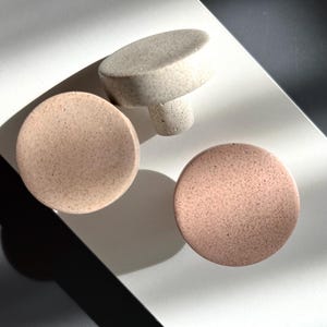 Puede incluir: Tres pomos de cer&aacute;mica moteada en tonos beige y rosa. Dos son redondos y uno tiene forma de hongo. Los pomos est&aacute;n dispuestos sobre una superficie blanca con una sombra oscura.