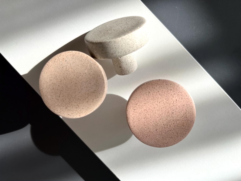 Puede incluir: Tres pomos de cer&aacute;mica moteada en tonos beige y rosa. Dos son redondos y uno tiene forma de hongo. Los pomos est&aacute;n dispuestos sobre una superficie blanca con una sombra oscura.