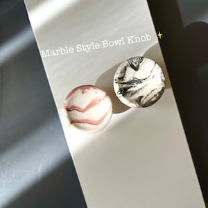 May include: Two marble-style bowl knobs. One is white with pink veining, the other is white with black veining. The text "Marble Style Bowl Knob" is above the knobs. The knobs are on a white surface.