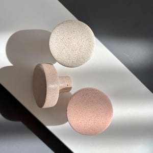 Puede incluir: Tres pomos de armario redondos y moteados en tonos neutros. Un pomo es de color beige claro y los otros dos son de color rosa p&aacute;lido. Un pomo rosa tiene un v&aacute;stago cil&iacute;ndrico. Los pomos est&aacute;n dispuestos sobre una superficie blanca y negra.